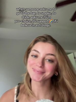 A young woman with long blonde hair, wearing a white and black patterned top and gold hoop earrings, looks towards the camera smiling. Text in a speech bubble is at the top of the screen.