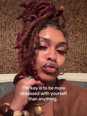 A selfie showing a woman with dark skin. She has a nose ring and large gold hoop earrings, with auburn dreadlocks styled up. She is wearing a brown top, and the background is a woven texture in shades of brown. The image has text overlay.