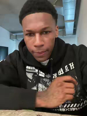 A medium shot shows a young Black man in a black hoodie, looking at the camera. The hoodie has a graphic on it. The background shows a metallic overhead structure.