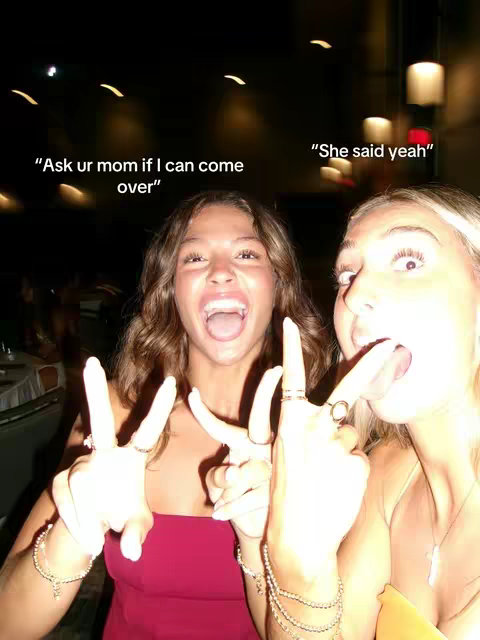 Two young women are smiling broadly, holding up peace signs with their fingers. The background is blurred, suggesting a social setting, possibly a party or gathering.