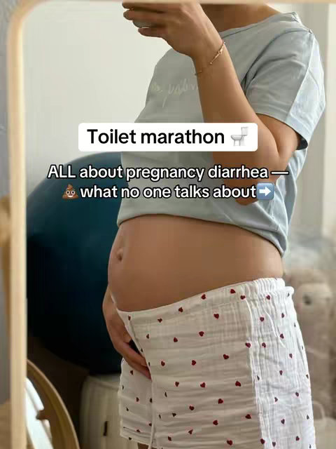 A woman is visible in a mirror, focused from the waist up. She appears to be pregnant, with a visible baby bump. She wears a short-sleeved light blue t-shirt and white shorts patterned with small red hearts. The image is framed by a mirror, with elements of a room visible in the background, including a door frame and a blue exercise ball creating a sense of a home setting. Text overlays on the mirror detail the video's focus on pregnancy issues.