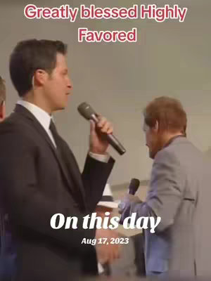 The image is a vertical video still showing two men in a church setting. The man on the left is centered, wearing a dark suit and tie, holding a microphone and speaking. The man on the right is further back, wearing a light gray suit jacket, also holding a microphone, facing away from the camera. Above them is a text overlay in a pinkish-red color. Underneath the men is a white text overlay with the date on the bottom.