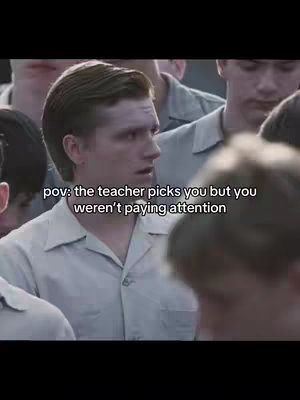 The image shows a still from a movie, focusing on the face of a young man. He has light skin, short, styled brown hair, and is wearing a light-colored collared shirt. His expression is one of surprise or slight alarm. Behind him are other young men, also in similar attire, suggesting a school or institutional setting. The lighting seems natural, with a focus on the main subject. The background is slightly blurred, which highlights the man's face. Text overlay is present, indicating the context of the scene.