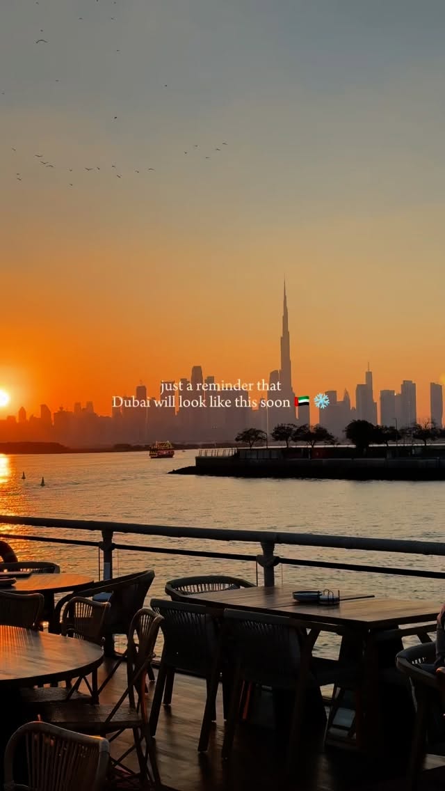 An eye-level shot of a sunset over a body of water, possibly a bay or harbor. Silhouetted buildings of Dubai are in the distance along with the Burj Khalifa. The sky is a gradient of colors, transitioning from a bright orange near the horizon to a softer blue higher up. A railing and some chairs are visible at the bottom, suggesting a waterfront restaurant or seating area.