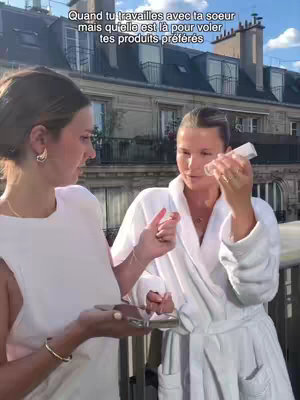 Two women are on a balcony, one is wearing a white robe and holding a skincare product. The other wears a white sleeveless top and is holding a small makeup product over a mirror. The background shows buildings and sky.