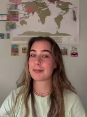 A young woman with light brown hair and fair skin smiles at the camera in front of a world map and multi-colored posters. She's wearing a light green shirt, and a delicate gold necklace is visible.