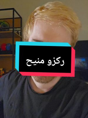 A selfie-style shot with a medium shot of a man with a light complexion and light blonde combed-back hair. The man is wearing a black t-shirt. His face is partially obscured by a rectangular overlay that is blue, with the exception of a central red rectangle. The background is slightly blurred, but appears to be a room with neutral walls and an indistinct piece of furniture.