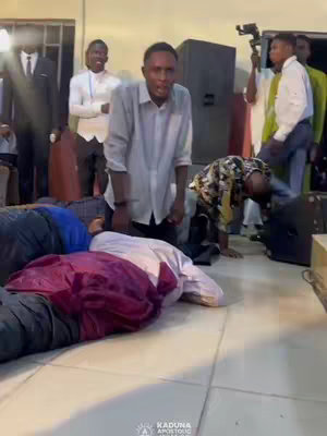 A group of people are gathered in a room, with some lying prostrate on the floor, and others standing. A man in a light gray shirt is positioned in the center, looking towards the camera with an open mouth, as though he is speaking emphatically. Several other figures, including some appearing to be dressed in suits, are around the room, giving an impression of a religious or spiritual event. The background includes a wall and what appears to be electronic equipment, accompanied by a logo in the lower-left corner. Overall, the setting suggests a place of worship or a similar gathering space with moderately lit conditions.