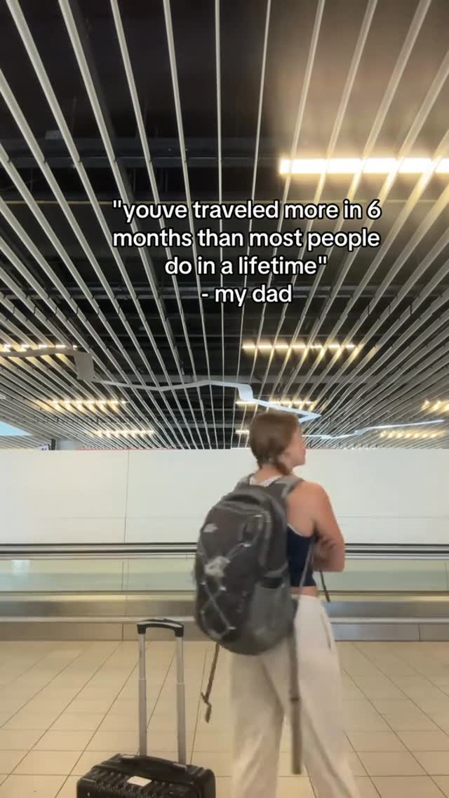 A person with short hair is seen from the back, wearing a dark tank top, light pants, and carrying a backpack, standing in a brightly lit airport or train station. A suitcase with a pull-up handle is to the person's left. Overhead, there are parallel lines of silver panels. Text overlays at the top of the image read: 'youve traveled more in 6 months than most people do in a lifetime" - my dad'.