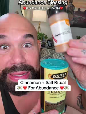The cover image features a close-up of a man's face with wide eyes and a smiling expression. He appears to be the main subject, holding a container in front of him. The top section of the image has a text overlay 'Abundance Blessing (For the 1st Of Every Month)'. The lower portion of the image, there is a box with text 'Cinnamon + Salt Ritual For Abundance'. The background is blurred, with elements of a room visible.