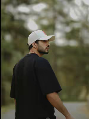 A man wearing a white baseball cap and a black t-shirt is walking, with a blurred green background, suggesting an outdoor setting. The shot is from the side, capturing his profile.