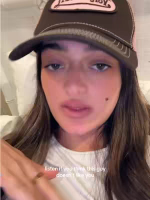 A young woman with long brown hair is wearing a baseball cap and looking directly at the camera. She is seated indoors, appearing to be in a casual setting, possibly a bed or a couch. The lighting seems to be soft, and the overall composition is a close-up, focusing on her face.