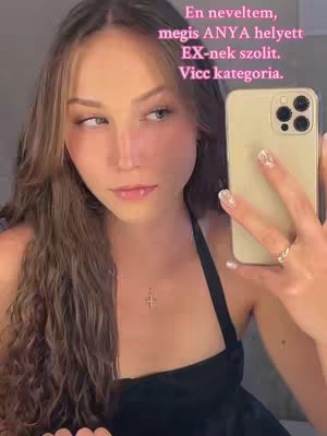 A young woman with long wavy brown hair is taking a selfie in a bathroom. She's holding a gold iPhone and wearing a black top and a necklace with a small cross pendant. There's text overlayed in the image in pink.