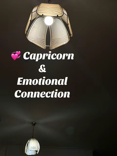 The image shows a dark room with two hanging lamps and a text overlay. The lamps have unique geometric shapes and are lit, creating a warm glow against the dark background. The text overlay consists of the words "Capricorn & Emotional Connection" in a bold white font, with pink heart emojis before the word "Capricorn". The composition is simple, focusing on the text and atmospheric lighting of the lamps.