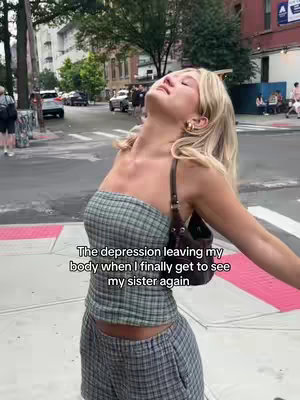 A woman stands outdoors with her head tilted back and arms outstretched, appearing joyful. She wears a plaid tube top and shorts, with a small purse on her shoulder. The background shows a street scene with buildings, cars, and other people.