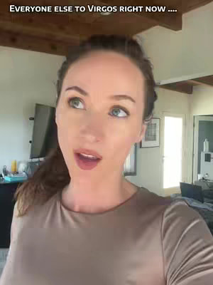 A woman with fair skin and light brown hair is positioned indoors. She looks upwards slightly, speaking with her mouth slightly open. The background shows a room with wooden beams, a window, and minimal decorations.