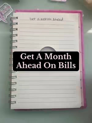 The cover image shows a notebook open, with a pink cover and spiral binding, on what looks like a light green surface. The lined pages of the notebook have text at the top that says "get a month Ahead". A black rectangular text overlay across the middle of the open pages reads "Get A Month Ahead On Bills". The background is neutral, with a small sticker on the upper left that looks like a printer.