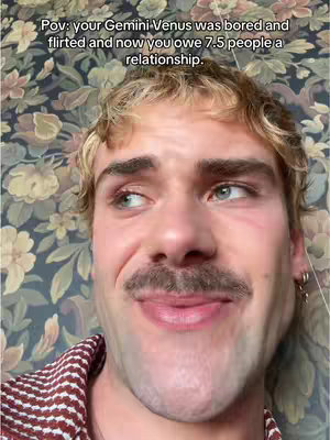 A close-up shot featuring a man with a mustache, looking upwards with a slightly mischievous expression, set against a busy floral wallpaper background. The man is wearing a checkered shirt.