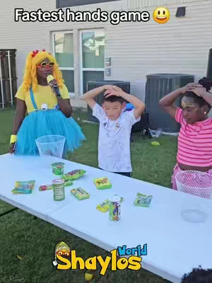 A group of three children and an entertainer are positioned behind a long white table, presumably outdoors. The central figure is a young boy, raising his hands towards his head. To his right, a girl also raises her hands. The entertainer, a woman with yellow hair, a yellow top, and a blue skirt, holds a microphone. Various food items and small baskets sit atop the table. The background includes a house, some air conditioning units, and green grass. The words "Shaylos World" are visible at the bottom of the image. 