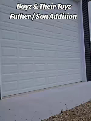 The cover image shows a white garage door with the words "Boyz & Their Toyz Father / Son Addition" overlaid on it. The door takes up most of the frame, with a cement walkway at the bottom and black siding on the right side. The image has a clean, bright look with a focus on the white garage door.