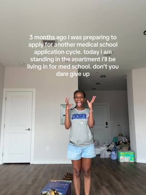 A woman with dark skin stands in a well-lit, sparsely furnished apartment. She wears a grey t-shirt, denim shorts, and has her arms raised, seemingly in excitement or disbelief. Text overlays the image, describing her journey toward medical school.