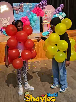 Two young people stand facing each other, each with balloons attached to their bodies, giving the impression of a game or activity. The background features a decorated space with pink and blue balloon arrangements. One person has a red balloon 'dress' and the other has a yellow balloon 'dress'.