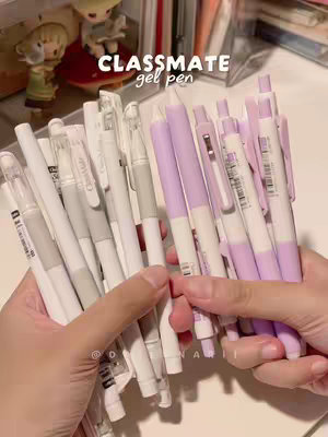 The image showcases a close-up view of a pair of hands holding a collection of purple and white gel pens, along with the text "CLASSMATE gel pen." The pens are the focal point, with a diverse mix of white and various shades of purple. The background seems to be a desk setting. A small figurine is also visible, along with a few books.