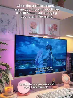 A well-lit, cozy workspace setup with a large monitor displaying an anime scene. Above the monitor, there are framed pictures. The desk includes a keyboard, a lamp, and decorative items such as a candle and a decorative light in the foreground. A quote from the anime is overlaid on the image.