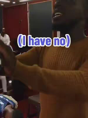 A man with dark skin is shown from the chest up, singing with his mouth open. A speech bubble with white text and a pink background is added at the center of the image. The background is blurry, which appears to be a recording studio, with other people.