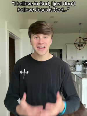 A young man with brown hair is centered in the image, speaking to the camera with an open mouth and raised hands. He wears a black long-sleeve shirt with a white cross graphic. Above him, text overlays read, "I believe in God, I just don't believe Jesus is God…" The background shows a well-lit domestic interior, including a white door, kitchen cabinets and a small chandelier.