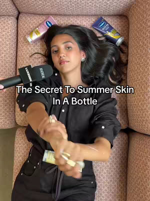 A woman is lying on her back on a pink couch, angled to the camera. Her dark hair fans out. She holds a black microphone in her left hand and is applying lotion to her right. Three product tubes are strategically placed as well.
