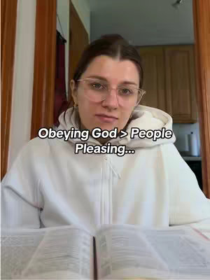 A woman wearing glasses and a white hoodie is seated, looking directly at the camera with a slightly stoic expression. The image shows a partially open book in front of her, implying she is reading or studying. The background is partially visible and includes a wooden door frame as well as a glimpse of a kitchen.