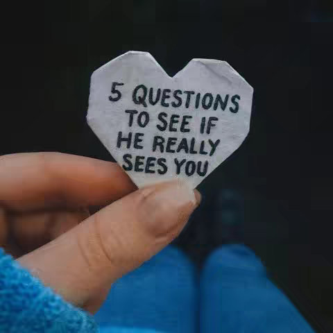 A close-up shot shows a hand holding a white paper heart with black text. The heart is slightly crumpled, and the text reads, '5 QUESTIONS TO SEE IF HE REALLY SEES YOU.' The background is a dark, blurred outdoor setting. The hand is wearing a blue sleeve, and blue jeans are visible below.