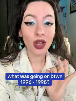 A woman with long dark hair, heavy eye makeup, and a cream-colored blouse is centered in a medium shot. A blue rectangle with white text overlays the bottom of the image. She is wearing turquoise earrings, and the background is out of focus. Her skin tone is light, and she has a neutral expression with her mouth slightly open. Her makeup looks bold, particularly the dark eyeliner. The overall composition is focused on her face and upper body.