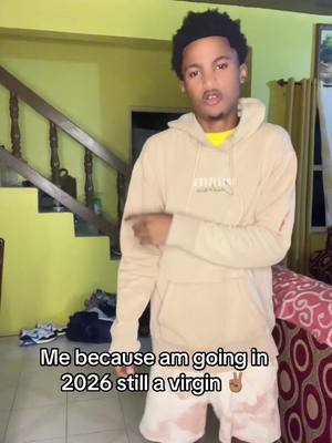 A young man in a beige hoodie stands in a room and points. Text at the bottom reads, 'Me because I'm going in 2026 still a virgin.' Stairs and a couch are visible in the background.