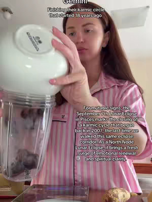 A woman with long brown hair is seen from the chest up, wearing a light pink striped pajama top, pouring ingredients from a white bowl into a clear blender. The background is a kitchen setting with a neutral color palette.