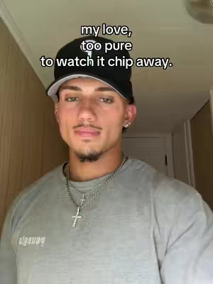 A young man in a grey t-shirt and a black cap with textual overlays. He has a silver necklace with a cross pendant, and the background seems like part of a house setting.