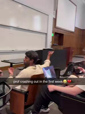 The cover image captures a lecture hall scene. Seated in the foreground, a man with dark hair gestures emphatically with his hands, appearing animated. A laptop screen is visible in front of him. The background features a white whiteboard, a projection screen, and a glimpse of the hall setting. The color palette is generally muted, with the man wearing clothes in lighter tones contrasting with the darker surroundings. The overall composition indicates a moment of surprise or intensity.