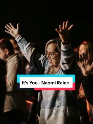 An image of a woman with blonde hair, wearing glasses and a sweater is singing worship. Her arms are raised in the air. There are other people in the blurred background, with one man facing away. A text overlay reads It's You - Naomi Raine.