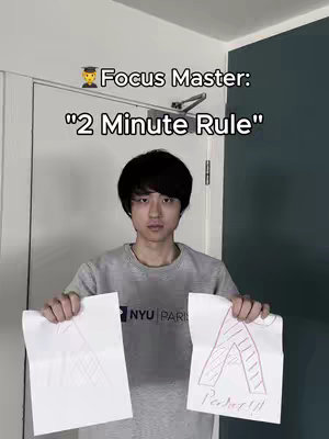 A young man stands in front of a white door and a teal wall, holding two torn pieces of paper. Text overlays at the top read "Focus Master: "2 Minute Rule"". He is wearing a grey t-shirt. The paper in his left hand has rough sketch marks, and the paper in his right has an 'A' drawn, along with the word 'PERFECT!!!'.