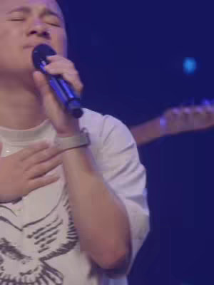 A person in a white t-shirt, holding a microphone, closes their eyes and touches their chest with their left hand. Behind, a blurry image of a band member on a guitar is visible. The stage is dimly lit with blue hues.