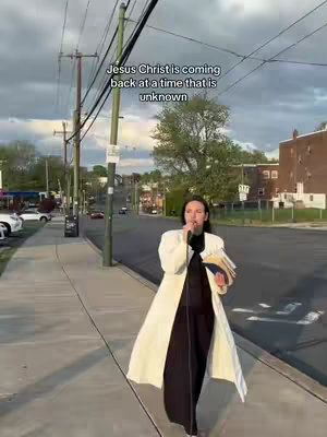A woman stands on a sidewalk, holding a microphone and some books, with a street and houses in the background. Text overlayed states 'Jesus Christ is coming back at a time that is unknown'.