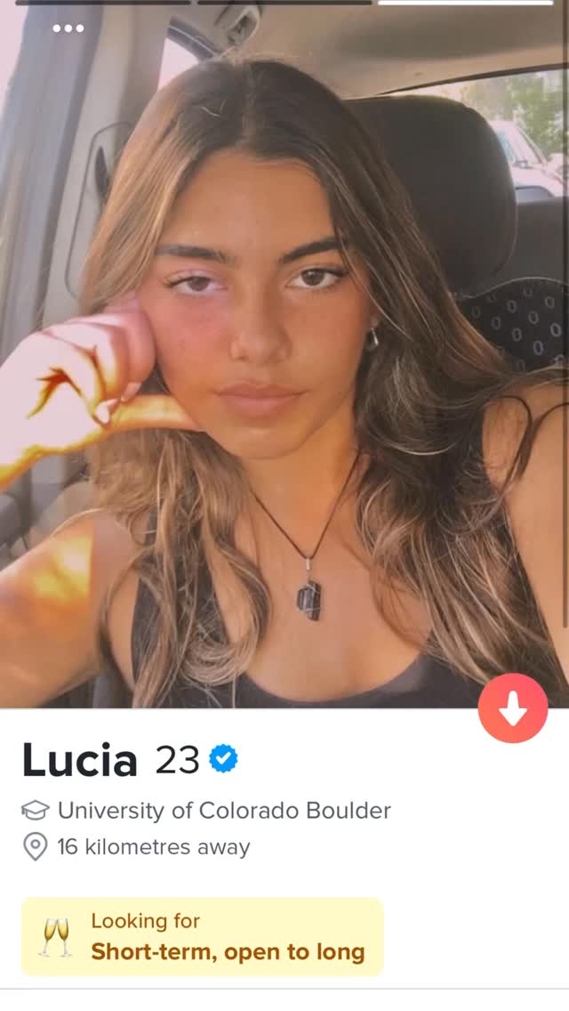The image is a screenshot from a dating app, featuring a young woman in a car setting. She has long hair and is looking at the camera with a slight expression. The app display includes her name, age, university, and distance from the viewer, along with a 'Looking for' and 'Short-term, open to long' description.