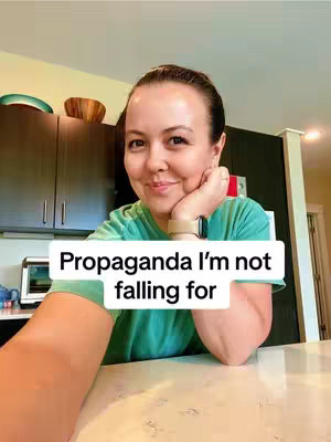 A woman with light skin, long dark hair, and a green shirt smiles brightly for a selfie. She rests her chin on her hand and the text overlay, reading "Propaganda I'm not falling for", is placed in front of her. The background suggests a kitchen setting with dark cabinets and appliances.