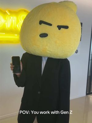 An image featuring a person dressed in a black suit, with a large yellow head shaped like a lemon character. The person is holding a black smartphone. The background is a blurred office-like setting. Text overlaid at the bottom says, "POV: You work with Gen Z."