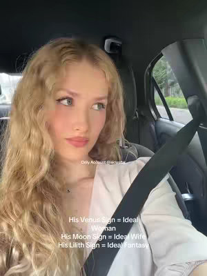A woman with long wavy blonde hair is sitting in a vehicle. She is wearing a white top and a seatbelt. Text overlays are present in the image.