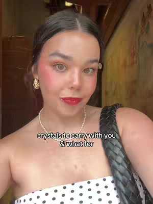 A woman with dark hair, wearing a white top with black polka dots and a black strap, poses for the camera. She has rosy cheeks, red lipstick, and gold earrings. A text overlay reads: "crystals to carry with you & what for". The background shows a blurred, indoor setting with warm lighting.