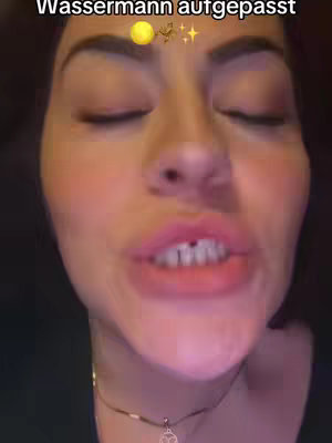 A close-up shot featuring a woman with dark hair. The focus is on her face, showing her eyes (closed), lips (slightly open, revealing teeth), and a small necklace. Above her head, there's text with a gold moon and stars. The lighting is soft, and the background is out of focus.