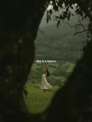 A vertical video image framed through a dark, blurred foreground of foliage. In the center, a slender woman in a white dress is captured mid-motion, her arms raised. The setting appears to be a green field with lush forest in the background, and the sky visible.