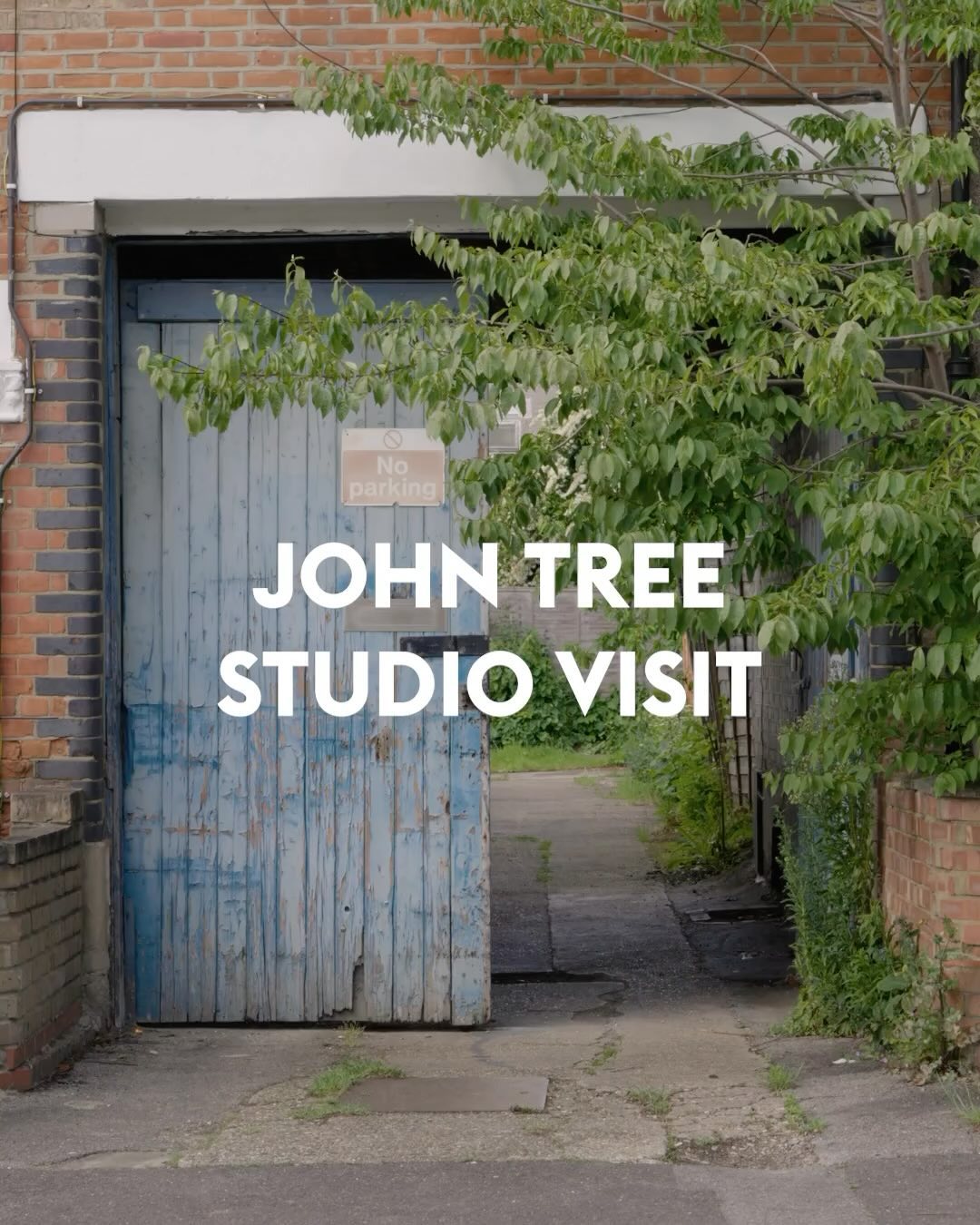 An outdoor shot revealing a blue wooden door slightly ajar, leading into a narrow alley. The door is weathered with peeling paint. The image is framed by brick and lush green foliage including a tree on the right side of the image. The text overlayed on the door reads “JOHN TREE STUDIO VISIT” in a bold white font.